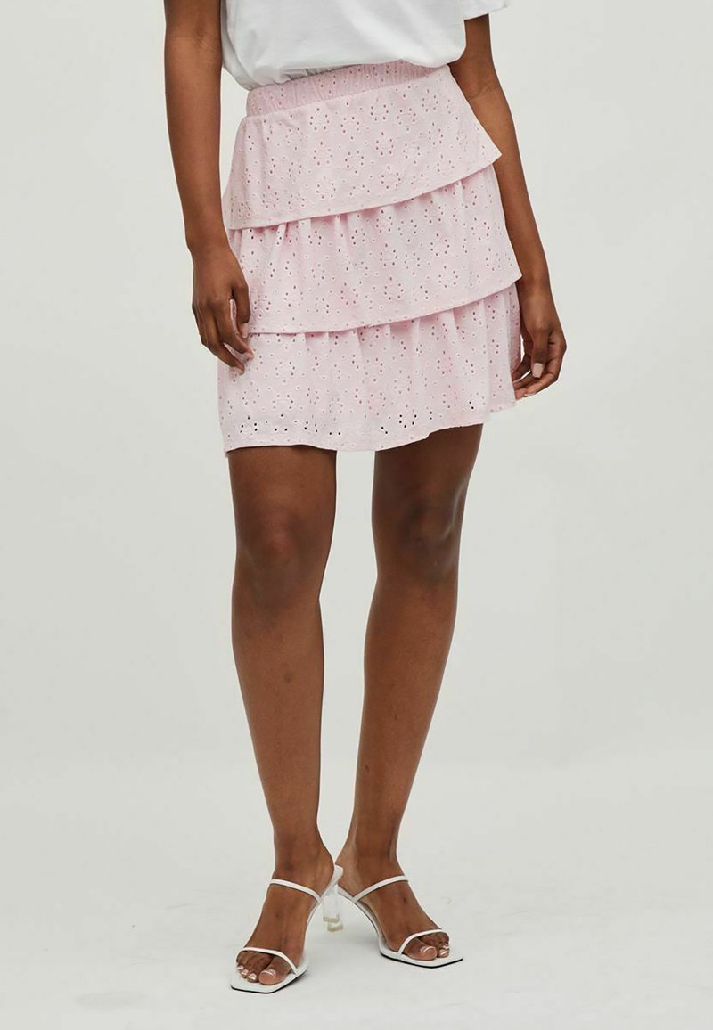 Jupe rose en couches avec un tissu à œillets, design en niveaux et taille élastique. Portée avec des sandales à talons transparents sur un fond neutre.