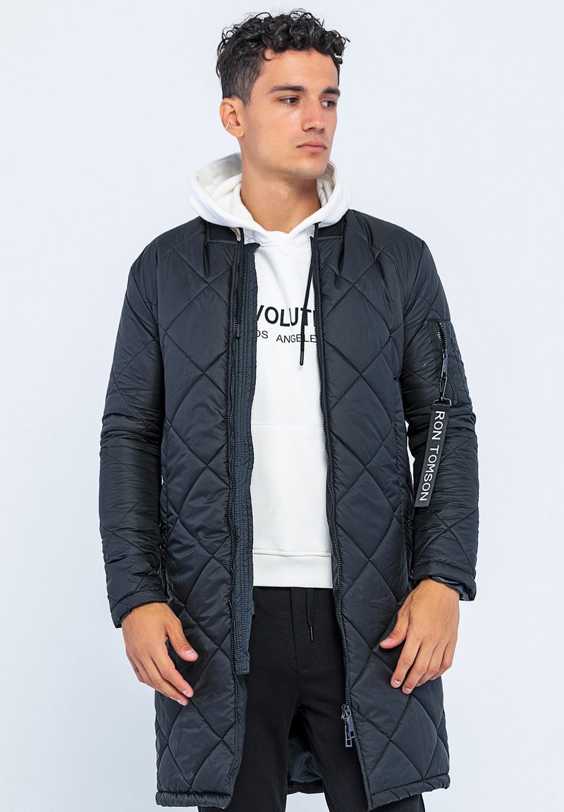 Zwarte gewatteerde lange jas met een rits aan de voorkant en zijzakken. Voorzien van een logolabel op de mouw en een gelaagd ontwerp over een witte hoodie.