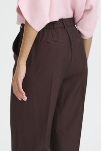 Frau trägt eine hochtaillierte, dunkelbraune, maßgeschneiderte Hose mit hinteren Leistentaschen und ein hellrosa, kurzärmeliges, in die Hose gestecktes Oberteil.