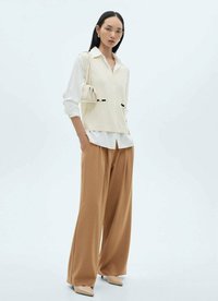 Ljusbeige ribbad ärmlös topp lager på lager över en vit skjorta, kombinerad med vida bruna byxor och komplementerad av en liten beige handväska.