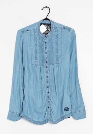 Hellblaues langärmeliges Jeanshemd mit durchgehender Knopfleiste, plissierten und verdrehten Steppnähten, hängend an einem schwarzen Kleiderbügel mit weißem Etikett.