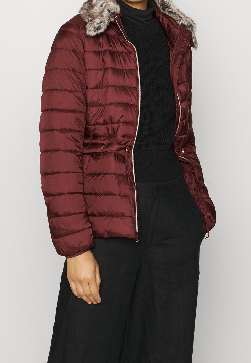 Femme portant une veste matelassée bordeaux avec un col en fausse fourrure, un pull en maille noir, et un pantalon noir à jambes larges, debout avec une main dans la poche.