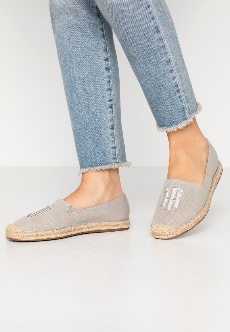 Personne portant un jean bleu clair à ourlet effiloché et des espadrilles beiges avec un logo brodé sur un sol blanc.
