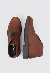 Botas de cuero marrón hasta el tobillo con superficie texturizada, diseño con cordones, puntera redondeada y suelas de goma negras en contraste.