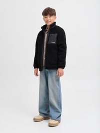 Black sherpa fleece jacket with a high collar, zippered chest pocket, paired with light blue wide-leg jeans and beige clogs.