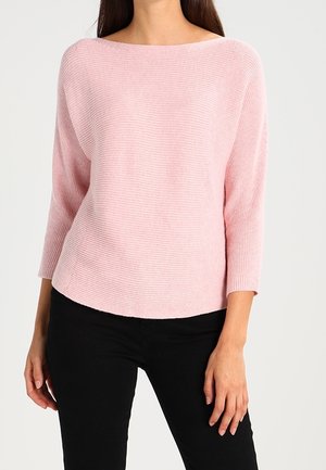 Frau trägt einen hellrosa gerippten Strickpullover mit Dreiviertelärmeln und schwarze Hosen vor einem schlichten Hintergrund.