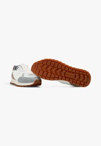Zapatillas blancas y grises con suelas de goma, una zapatilla en posición vertical y la otra mostrando el patrón de la suela y la marca "Scalpers".