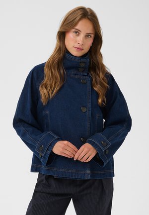 Femme aux longs cheveux bruns portant une veste en jean bleu foncé boutonnée et un pantalon foncé à fines rayures, sur un fond uni.