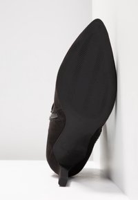 Zapato negro de ante con punta afilada y suela de goma texturizada negra, con un tacón delgado y un diseño lateral minimalista.