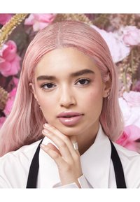 Jeune femme aux longs cheveux roses, portant une chemise blanche. Lèvres brillantes et roses et une bague en argent, avec un fond floral dans des tons doux de rose et de vert.