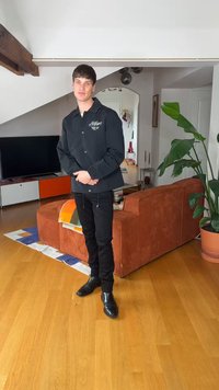 Νέος άνδρας με μαύρο μπουφάν και παντελόνι στέκεται σε ένα μοντέρνο σαλόνι με πορτοκαλί καναπέ, ξύλινο δάπεδο, μεγάλο φυτό και κονσόλα τηλεόρασης.