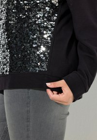 Felpa nera con un pannello di paillettes caratterizzato da paillettes circolari argentate. La felpa ha una vestibilità comoda, con pantaloni in denim grigi parzialmente visibili.