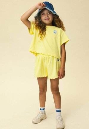 Child with curly hair wearing yellow scalloped shorts and top, blue bucket hat, white sneakers, and striped socks, standing against beige background.