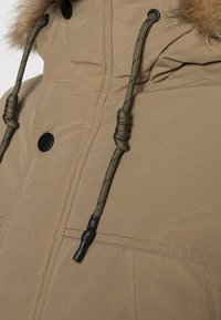 Detailní záběr hnědého zimního kabátu s černým patentním knoflíkem a zelenými stahovacími šňůrkami, s okolím kapuce podšitým kožešinou.