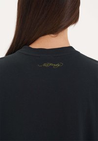 Zwarte t-shirt gezien vanaf de achterkant, met een ronde hals en een klein geel cursief logo op de bovenrug gedrukt. Zachte stoftextuur.