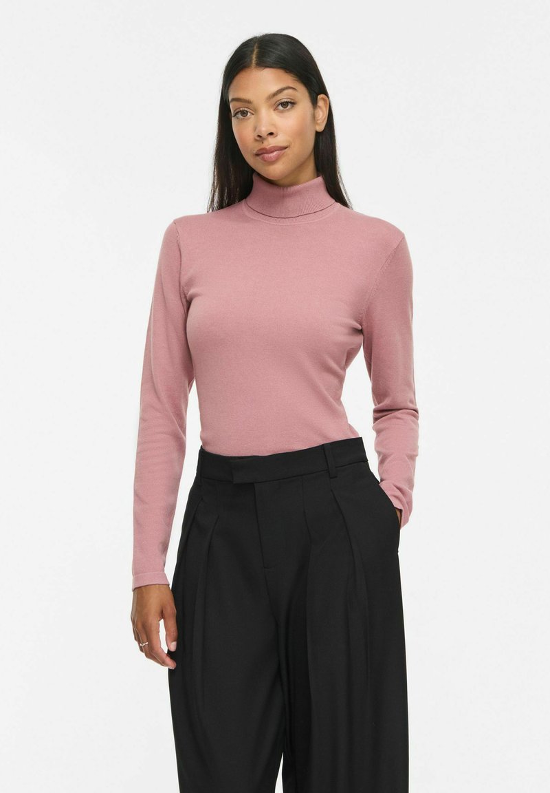 Femme aux longs cheveux foncés portant un pull à col roulé rose et un pantalon plissé noir taille haute, debout avec une main dans la poche.