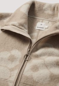 Beige en witte fleecejas met een hoge kraag, ronde ritssluiting en een "hessnatur" label binnenin de kraag.