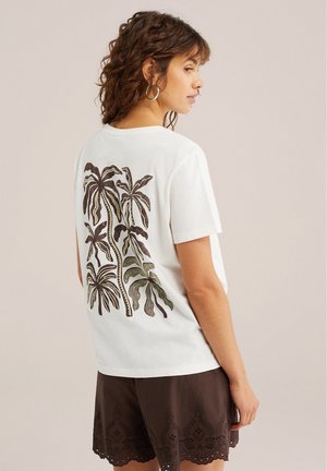 Mujer con pendientes de aro, vistiendo una camiseta blanca con estampado de palmeras en la espalda y pantalones cortos marrones con borde ondulado, vista de perfil.