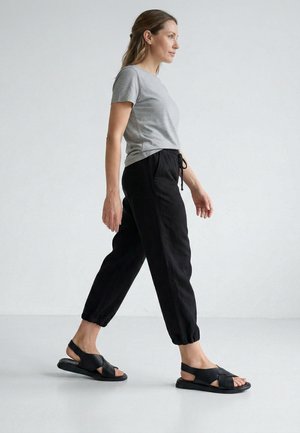 Kvinde går iført en grå kortærmet skjorte, sorte bukser med snøre og sorte åbne tå-slingback-sandaler på en ensfarvet lys baggrund.