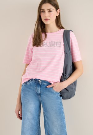 Jeune femme portant un t-shirt rayé rose avec le mot "Bonjour", un jean bleu clair, et un sac à bandoulière gris, devant un fond uni.