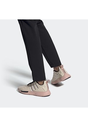 Ben iført sorte bukser og beige Adidas-sneakers med pink såler, der går på en lysegrå overflade.