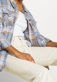 Persona con pantaloni beige, top bianco e camicia a quadri blu-marrone con bottoni, seduta con la mano appoggiata sulla coscia.