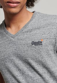 T-shirt grigio a maniche corte con scollo a V in tessuto strutturato. Presenta un piccolo logo blu "SuperDry" sul petto, che mette in risalto il suo design.