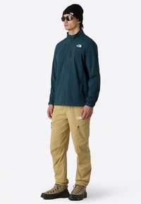 Ciemnoturkusowa kurtka zapinana na zamek z wysokim kołnierzem, beżowe spodnie, okulary przeciwsłoneczne i czarna czapka typu beanie. Charakteryzuje się akcentami z logo i dopasowanym krojem.