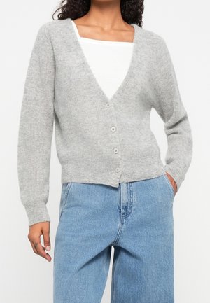 Frau trägt eine hellgraue, geknöpfte Strickjacke über einem weißen Top mit hellblauen High-Waist-Jeans, Hände entspannt an den Seiten.