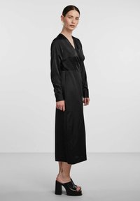 Black satin dress with long sleeves, V-neckline, and a fitted waist. The design is ankle-length, complemented by block-heeled black shoes.