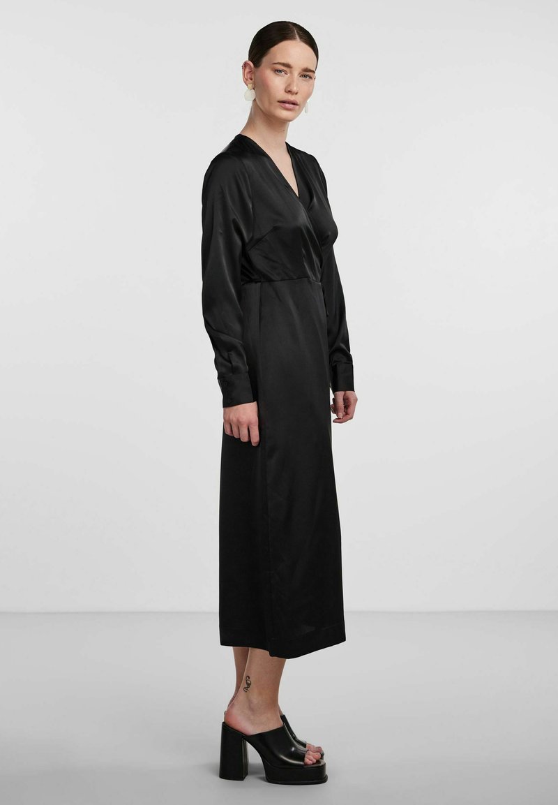 Black satin dress with long sleeves, V-neckline, and a fitted waist. The design is ankle-length, complemented by block-heeled black shoes.