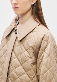 Μπεζ μπουφάν με επένδυση, φαρδιά γιακά, με μεγάλο κουμπί κλεισίματος και υφή διαμαντ�ένιου μοτίβου σε όλο το ύφασμα.