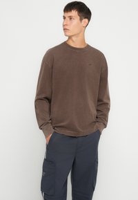 Hnedá mikina s dlhým rukávom, okrúhlym výstrihom a jemným logom. Tkanina sa zdá byť mäkká s jemne opotrebovanou textúrou.