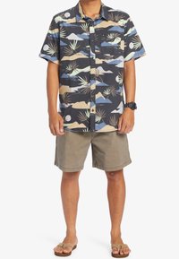 Koszula z krótkim rękawem z wielokolorowym, abstrakcyjnym wzorem na ciemnym tle, zestawiona z beżowymi szortami i brązowymi klapkami.