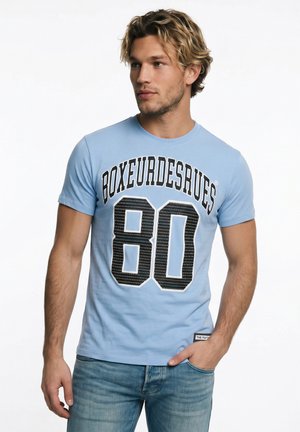 Junger Mann mit welligem blondem Haar, der ein hellblaues T-Shirt mit "BOXEURDESRUES 80" und blaue Jeans trägt, steht mit einer Hand in der Tasche.