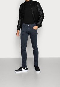 Hombre con chaqueta bomber de manga de cuero negro, cuello de tortuga negro, pantalones gris oscuro y zapatillas Nike en blanco y negro de pie sobre un suelo gris.