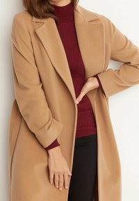 Kamelfärgad ullrock med spetsig krage, sidofickor och vida ärmar, i kombination med en vinröd rippad polotröja.