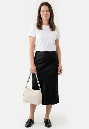 Nainen, joka pukeutuu valkoiseen lyhythihaiseen paitaan ja mustaan midihameeseen, kantaa beigeä käsilaukkua ja käyttää mustia loafereita, seisoo valkoisen taustan edessä.