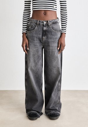 Person iført grå jeans med høj talje og løs pasform med frynsekanter, kombineret med sorte sneakers og en sort-hvid stribet cropped langærmet top.