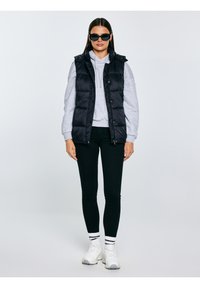 Naine, kellel on mustad päikeseprillid, must suletud jope halli kapuutsiga, mustad retuusid, valged sokid ja valged tossud, seisab valgel taustal.