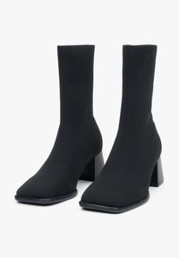Czarne botki na wysokim obcasie wykonane z teksturowanej tkaniny, z kwadratowym czubkiem i masywnym słupkowym obcasem. Bezszwowy design bez widocznych zapięć.