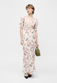 Kvinne i hvit blomstrete maxikjole med blonder, holder en liten grønn veske, iført svarte spisse sko, stående på hvit bakgrunn.