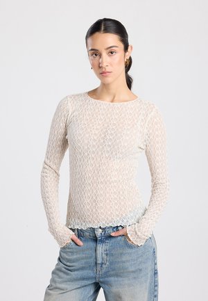 Frau mit dunklem Haar in einem niedrigen Pferdeschwanz, trägt ein weißes langärmeliges Spitzentop und blaue Jeans, steht mit den Händen in den Taschen vor einem schlichten Hintergrund.