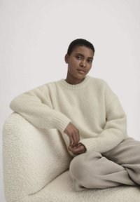 Witte gebreide trui met een ronde hals en geribbelde mouwen. Het model zit op een fluffy, neutraal gekleurde stoel, met beige broek aan.