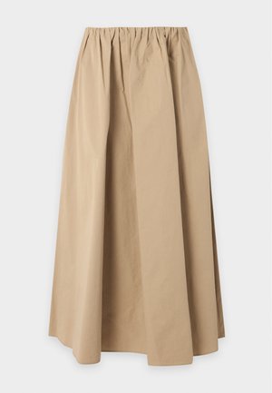Femme portant une jupe longue beige, un haut rayé à manches longues, des chaussures marron à glands et tenant un petit sac beige avec une chaîne.