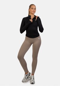 Crna majica s dugim rukavima i ovratnikom s polu-zipom, uparena s svijetlosmešavim tajicama s glatkom teksturom i naglašenim logom. Bijele tenisice upotpunjuju izgled.