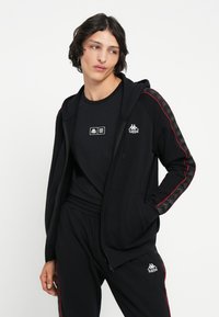 Sudadera negra con cremallera y logo, detalles en rojo y mangas estampadas; combinada con una camiseta negra y pantalones a juego con franjas laterales.