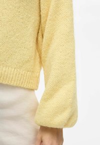 Rumena pletena jopica z teksturiranim zaključkom, ohlapnimi rokavi in ribastim robom. Mehka tkanina vidna v bližnjem pogledu.