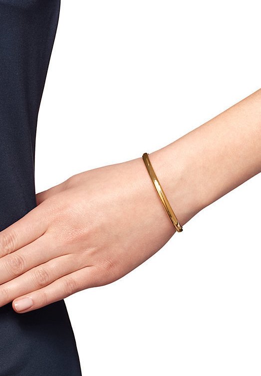 Gold bangle bracelet on a wrist, featuring a smooth, polished finish with a slender, rounded design, resting against a dark fabric background.