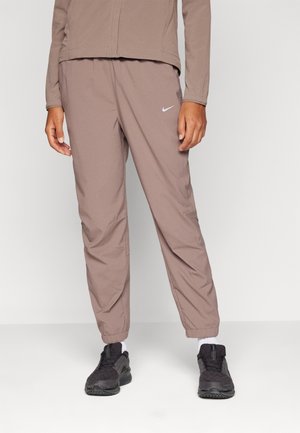 Person trägt mauvefarbene Nike Trainigsanzughose mit elastischen Bündchen, schwarze Sneaker, weiße Socken und passendes mauvefarbenes Langarmoberteil vor einfarbigem Hintergrund.
